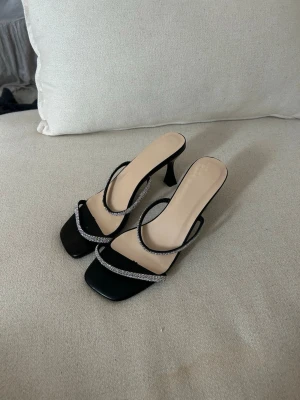 Svarta sandaler med strass och klack - Säljer ett par svarta sandaler med fyrkantig tå och smala remmar täckta av glittrande strass. Skorna har en elegant låg klack och öppen tå, perfekta för att lyfta vilken outfit som helst. Materialet är syntet och de har en stilren, modern look.