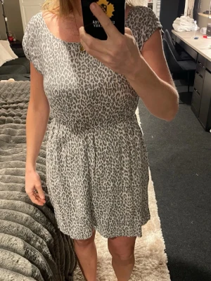 Grå leopardmönstrad klänning från ONLY - Säljer en kort klänning från ONLY i storlek 38 med grått leopardmönster. Klänningen har rund halsringning, korta ärmar och ett lätt rynkat midjeparti som ger en snygg siluett. Materialet känns mjukt och luftigt, perfekt för dig som gillar djurmönster.