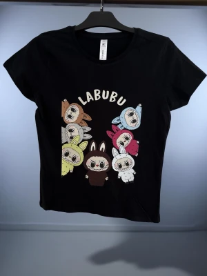 Svart LABUBU t-shirt med tryck - Svart t-shirt med färgglatt tryck av flera tecknade LABUBU-figurer på framsidan. Modellen är kortärmad med rund hals och har en normal passform. Materialet är mjukt och skönt, troligtvis bomull. Perfekt för dig som gillar unika och lekfulla motiv.