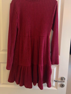 Vinröd ribbad kortklänning - Säljer en vinröd kortklänning med långa ärmar och ribbad struktur. Klänningen har en lätt utställd kjol och rund halsringning. Perfekt för dig som gillar en enkel men snygg stil med lite extra volym nertill.