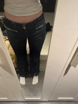 Mörkblå bootcut jeans med dragkedjor - Säljer ett par mörkblå jeans från Miss Sixty med bootcut passform och låg midja. Jeansen har dubbla knappar, dekorativa dragkedjor framtill och snygga broderade detaljer på bakfickorna. Klassisk jeanslook med en twist, perfekta till sneakers eller boots.