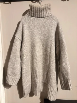 Grå stickad polotröja - Säljer en grå stickad polotröja med oversized passform och bred ribbad krage. Tröjan har långa ärmar och ribbade muddar vid ärmslut och nederkant. Perfekt för kyliga dagar och enkel att styla med jeans eller leggings.