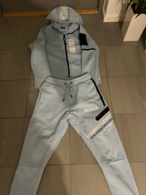 Ljusblå Hoodrich tracksuit set S - Säljer ett ljusblått tracksuit set från Hoodrich i storlek S. Setet består av en hoodie med dragkedja, huva och vita tryck samt matchande byxor med snörning och tryckdetaljer. Svarta detaljer på fickor och ärmar ger extra stil. Perfekt för chill eller streetwear.