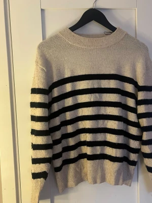 Randig stickad tröja från H&M - Säljer en beige stickad tröja från H&M i storlek S med svarta horisontella ränder. Tröjan har rund hals, ribbade muddar och långa ärmar. Perfekt för dig som gillar klassisk och chill stil.