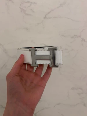 Vit bälte - Säljer ett stilrent vit stilrent bälte med de ikoniska H-loggan. Den ör helt ny aldrig använd.
