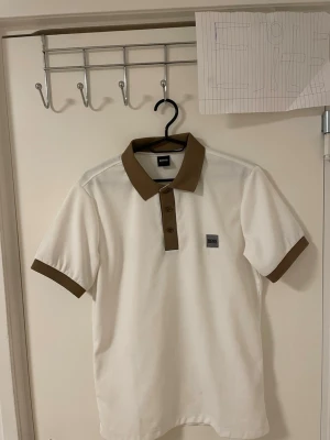 Vit och brun Pike BOSS - Säljer en stilren polotopp från BOSS i vit färg med bruna detaljer på krage, knappslå och ärmslut. Toppen har korta ärmar och en klassisk pikékrage med tre bruna knappar framtill. Perfekt för dig som gillar enkel och snygg design.Liknande modell som originala.