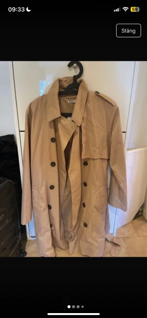 Beige trenchcoat från C, strl 40 - Säljer en beige trenchcoat från Cubus  i storlek 40. Jackan har klassisk dubbelknäppning med svarta knappar, axelklaffar och bälte i midjan. Långärmad och tillverkad i bomull, perfekt för vår och höst. Snygg och tidlös modell med coola detaljer.