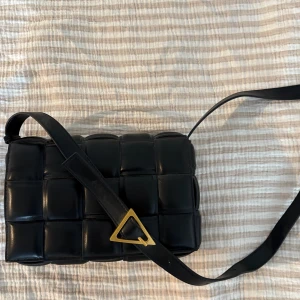 Svart väska  - Säljer en svart FEJK axelväska med puffad, quiltad design och bred axelrem med guldfärgat spänne i triangelform. Väskan är tillverkad i skinn och har ett innerfack med dragkedja. Perfekt för dig som gillar stilrena och lyxiga detaljer.