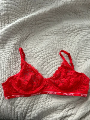 Röd spets-bh från Lounge - Säljer en röd balconette-bh från Lounge med bred resår och logga. Bh:n har tunna, justerbara axelband och är gjord i genombruten spets med fina detaljer. Materialet är 88% polyamid och 12% spandex för en stretchig och bekväm känsla.
