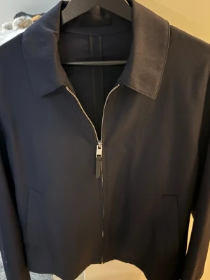 Mörkblå H&M jacka  - Snygg svart jacka från H&M i storlek S, med klassisk krage och silverfärgad dragkedja framtill. Jackan har två diskreta fickor på sidorna och är långärmad. Materialet känns mjukt och har en stilren look som passar till många outfits.