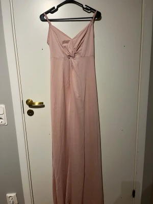 Ljusrosa långklänning med knut - Säljer en ljusrosa långklänning med smala axelband och snygg knutdetalj framtill. Klänningen har en v-ringad hals och ärmlös design, vilket ger en stilren och feminin look. Materialet känns lätt och mjukt mot huden, perfekt för varma dagar. Använd en gång, finns en liten fläck längst ner på klänningen men inget synligt 
