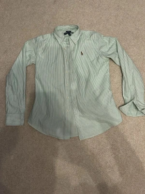 Grön- och vitrandig skjorta Ralph Lauren - Säljer en slim fit skjorta från Ralph Lauren i grön- och vitrandigt mönster. Skjortan har klassisk krage, knappar framtill och den ikoniska broderade loggan på bröstet. Tillverkad i mjuk bomull och har långa ärmar med manschett.