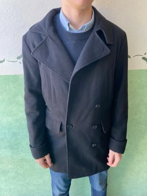 Svart skepparkavaj med bred krage - Klassisk svart skepparkavaj med dubbelknäppning och bred krage. Jackan har långa ärmar, två fickor framtill och är tillverkad i ett tjockare ullmaterial som håller dig varm under kyliga dagar. Perfekt för dig som gillar stilren och tidlös look.