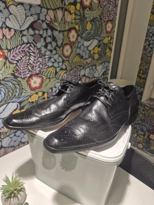 Svarta klassiska skinnskor med snörning 42 - Snygga svarta skinnskor med spetsig tå och klassisk broguemönstrad design. Skorna har snörning och låg klack, perfekta för dig som gillar stilrena detaljer. Materialet är äkta skinn och de har en platt sula med diskreta perforeringar för extra karaktär.