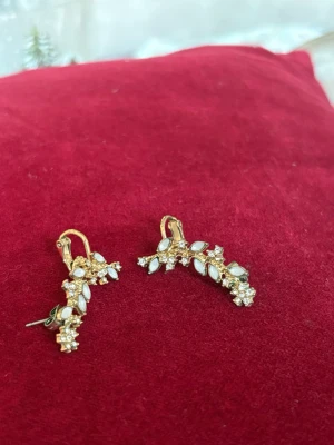 Guldiga örhängen med vita stenar - Säljer ett par snygga örhängen i guldton med vita bladformade stenar och små gnistrande kristaller. De har en unik, böjd form som följer örats kant och stängs med både krok och plopp. Perfekta för dig som vill ha något extra till din look.
