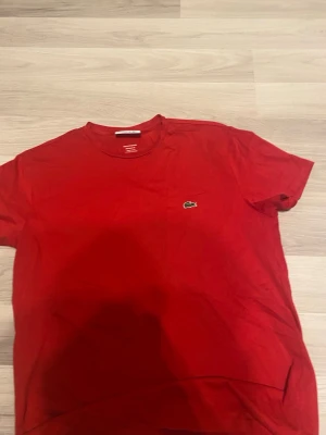 Röd t-shirt från Lacoste - Säljer en röd t-shirt från Lacoste med klassisk krokodillogga på bröstet. T-shirten har rund hals och korta ärmar, perfekt för en chill och stilren look. Materialet är mjuk bomull som känns skönt mot huden.