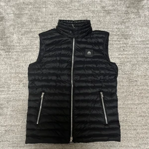  Moose Knuckles väst - Säljer en svart dunväst från Moose Knuckles med quiltad design och hög krage. Västen har två fickor med dragkedja och ett snyggt logomärke på bröstet. Perfekt för lager-på-lager och chill stil under kyliga dagar.