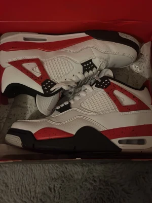 Nike Air Jordan 4 red cement - Nike Air Jordan 4 red cement sneakers med svarta och röda detaljer och den klassiska Jordan-loggan på plösen och yttersulan. Tillverkade i skinn och syntetmaterial, med platt sula och rund tå.