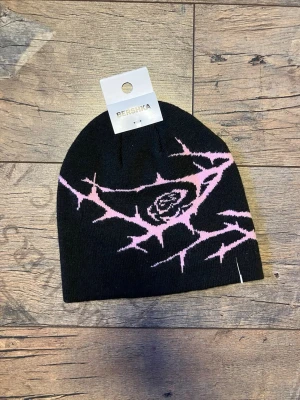 Helt ny swag beanie från Bershka - Helt ny med prislapp kvar! Svart/rosa mössa från Bershka med taggmönster och en ros i mitten. Stickad i mjukt syntetmaterial som sitter skönt på huvudet. Perfekt för dig som gillar edgy detaljer och vill sticka ut lite extra.
