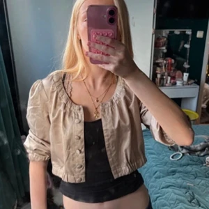 Beige Bolero från Zoul - Säljer en beige bolero från Zoul med rundad hals och puffiga långärmar. Jackan har knappar framtill och är tillverkad i ett lätt syntetmaterial. Perfekt för dig som vill ha en chill och trendig look.