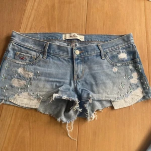 Lågmidjade ljusblå jeansshorts - Ljusblå jeansshorts från Hollister med råa kanter, slitningar och broderade detaljer med små pärlor framtill. Klassisk femficksmodell med låg midja och snyggt sliten look. Perfekta för sommaren och ger en avslappnad vibe.