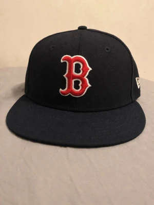 Svart Boston Red Sox 59FIFTY keps - Säljer en svart 59FIFTY keps från New Era med Boston Red Sox logga i rött och vitt framtill. Kepsen har en platt skärm och broderade MLB- och New Era-loggor på sidorna och bak. Tillverkad i polyester och har en klassisk, stilren look.