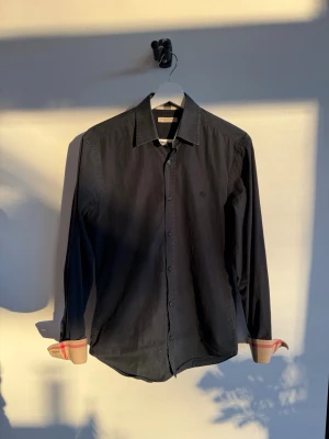 Svart Burberry skjorta med rutiga detaljer - Svart skjorta från Burberry med klassiska rutiga detaljer på insidan av krage och manschetter. Skjortan har lång ärm, knappar framtill och broderad Burberry-logga på bröstet. Materialet är mjuk bomull och passformen är normal. Stl S