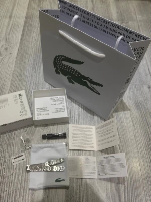 Silverfärgad Lacoste armbandslänk - Snygg armbandslänk från Lacoste i silverfärgad metall med stilrena rektangulära länkar och diskret Lacoste-logga. Kommer med originalpåse, box och verktyg för justering. Perfekt för dig som vill ha en clean och modern look.