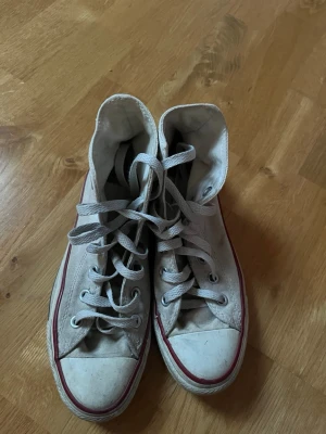 Vita Converse (höga) - Säljer ett par klassiska höga vita Converse med röd rand runt sulan. Skorna har snörning, rund tå och gummisula. Converse-loggan syns på sidan. Perfekta för dig som gillar retrostil.