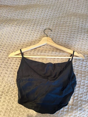 Svart festtopp i satin från Bershka - Snygg svart satinlinne från Bershka i storlek XS. Toppen har tunna justerbara axelband och ger en lyxig känsla. Perfekt att styla med jeans eller kjol för en trendig look.