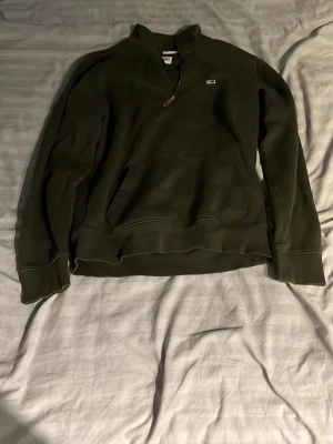 Tommy hilfiger fleece-tröja med half zip - Säljer en mörkgrön fleece-tröja från Tommy Hilfiger med half zip och lång ärm. Tröjan har en praktisk magficka och ett litet broderat logomärke på bröstet. Perfekt för kyliga dagar och enkel att matcha med jeans eller joggers. Storlek S