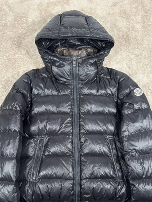 Svart Moncler pufferjacka med huva - Säljer en svart pufferjacka från Moncler med en glansig finish och huva. Jackan har två sidofickor med dragkedja, ribbade muddar och Moncler-logga på ärmen. Insidan är grå och jackan har quiltad design samt dragkedja framtill. Perfekt för kalla dagar.