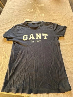 Svart GANT t-shirt med tryck - Svart t-shirt från GANT med stort vitt tryck på bröstet där det står 'GANT Est. 1949'. Klassisk rund hals och korta ärmar. Tillverkad i mjuk bomull som är skön mot huden. Perfekt för dig som gillar stilrena och sportiga plagg med tydlig logga.