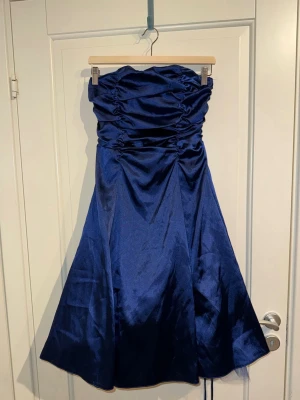 Marinblå axelbandslös klänning Juju & Christine - Snygg marinblå axelbandslös klänning från Juju & Christine med glansigt tyg och snörning i ryggen. Kjolen är utsvängd med voluminöst tyllfoder under för extra fluff. Den går ungefär till knäna.