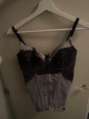 Grå korsett med svarta spetsdetaljer - Säljer en snygg grå korsett med svarta spetsdetaljer och svarta axelband. Framtill finns en liten rosett och pärlbeströdd X-detalj vid axelbandet. Korsetten har en figurnära passform och dragkedja baktill. Materialet är glansigt och känns lyxigt. Passar mig med storlek 36/75B , hör av er om ni vill ha bättre bilder💞