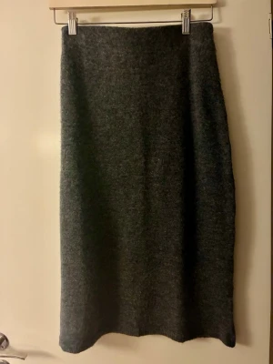 Stickad kjol från Mango  - Mysig mörkgrå kjol från MNG. Kjolen är tillverkad i en mjuk blandning av polyester, akryl och ull som ger en skön känsla och lite struktur. Perfekt att styla med chunky boots eller sneakers. Kjolen är ca 70 cm lång. Använd endast 2 ggr. 