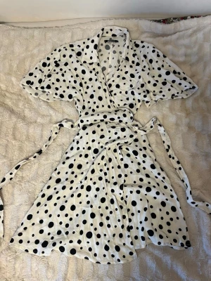 Prickig omlottklänning Gina Tricot  - Snygg vit omlottklänning från Gina Tricot i storlek XS med svarta prickar. Klänningen har korta ärmar, krage och knytband i midjan. Perfekt för dig som gillar lekfulla mönster och en feminin siluett. Endast använd en gång 