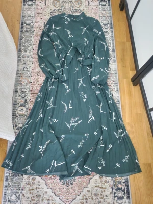 Grön långklänning med blommigt mönster - Grön långklänning med vitt blommigt mönster och knyt i midjan. Klänningen har långa ärmar och en rund halsringning. Materialet känns lätt och luftigt, perfekt för vår och sommar. 