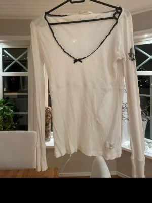 Vit ribbad topp Odd Molly - Vit ribbad långärmad topp från Odd Molly, modell 'memories top'. Toppen har svart spetskant och liten rosett vid v-ringningen samt ett tryckt motiv på ena ärmen. Tillverkad i mjuk bomull och har en figurnära passform. Trendig och smickrande topp! 