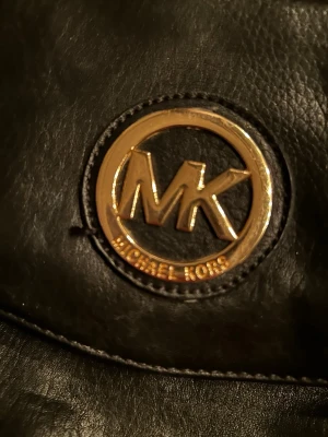 Svart kuvertväska Michael Kors - Snygg svart kuvertväska från Michael Kors i skinn med guldigt MK-emblem på framsidan. Väskan har dragkedja på baksidan och insidan, samt magnetknäppning. Perfekt storlek för det viktigaste och stilren design som lyfter din outfit.