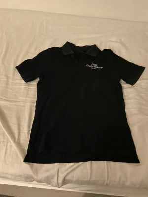 Svart Peak Performance piké - Säljer en svart pikétröja från Peak Performance med klassisk krage och knappslå framtill. Diskret logga broderad på bröstet och snygga detaljer på knapparna. Perfekt för en clean och sportig look.
