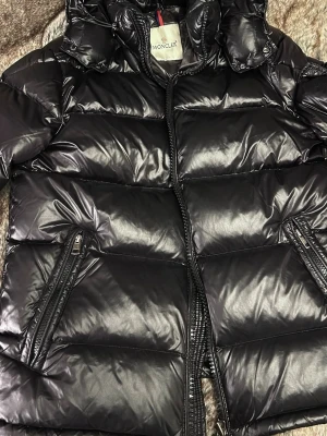 Svart Moncler pufferjacka med huva - Säljer en svart pufferjacka från Moncler med glansig finish och huva. Jackan har två sidofickor med dragkedja och tydliga Moncler-loggor på insidan. Materialet är polyester och dun, perfekt för kalla dagar. Stängs med dragkedja framtill.