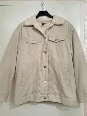 Beige teddyfodrad jeansjacka - Beige jeansjacka med mjukt teddyfoder och krage. Jackan har klassisk knappstängning, två bröstfickor med lock och sidofickor. Perfekt för dig som gillar chill och stilren look med extra värme. Köpt ett par år sedan så säljs inte längre i butik. 