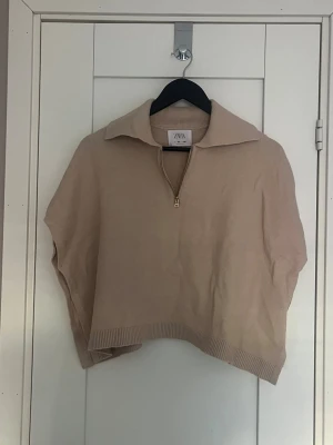 Beige stickad half zip tröja från Zara - Snygg beige stickad tröja från Zara med krage och half zip-dragkedja i guld. Modellen är croppad och har ribbade detaljer vid nederkant. Perfekt för dig som gillar en chill och trendig look.