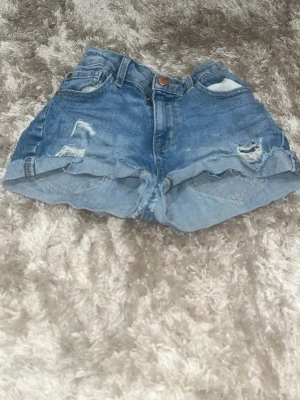 Blå denimshorts från Detroit - Säljer ett par blå denimshorts från Detroit med hög midja och slitna detaljer framtill. Shortsen har klassiska fem fickor, knapp och dragkedja samt uppvikta benslut. Perfekta för varma dagar och en chill stil.