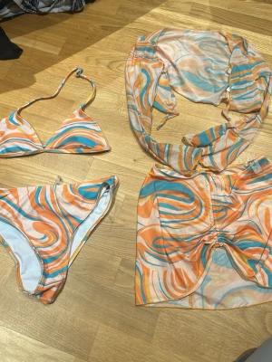 Färgglad mönstrad bikini från SHEIN 4 delat set - Säljer en bikini från SHEIN med swirl-mönster i orange, blått, gult och vitt. Överdelen har snygg rynkning framtill och båda delarna är gjorda i mjuk polyester med lite stretch. Perfekt för dig som gillar färg och coola mönster på stranden.