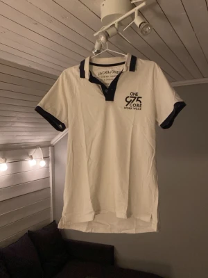 Vit piké från Jack & Jones - Säljer en vit pikétröja från Jack & Jones med svart krage och svarta detaljer vid ärmslut. Tryck på bröstet med texten 'ONE 975 CORE WORK WEAR'. Klassisk kortärmad modell i mjuk bomull, perfekt för en chill och stilren look.