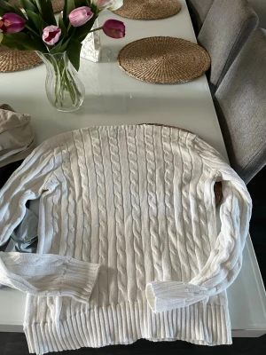 Vit kabelstickad tröja Polo Ralph Lauren - Vit kabelstickad tröja från Polo Ralph Lauren i storlek M. Tröjan har rund hals, långa ärmar och klassisk blå broderad logga på bröstet. Ribbstickade muddar vid ärmslut och nederkant. Perfekt för dig som gillar stilren och tidlös design.