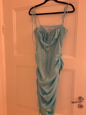 Blommig blå långklänning från Adoore, santorini - Säljer en ljusblå långklänning från Adoore med smala axelband och knytning fram. Klänningen har små gula blommor som mönster och snygga rynkade detaljer längs sidorna. Perfekt för sommarens alla tillfällen!