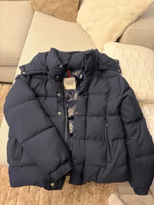 Mörkblå dunjacka från Moncler - Säljer en mörkblå dunjacka från Moncler med klassisk puffer-design. Jackan har hög krage, huva och dragkedja med tryckknappar. Moncler-logga på ärmen och flera praktiska fickor. Perfekt för kalla dagar och riktigt snygg till streetwear. NFC tagg finns på loggan.